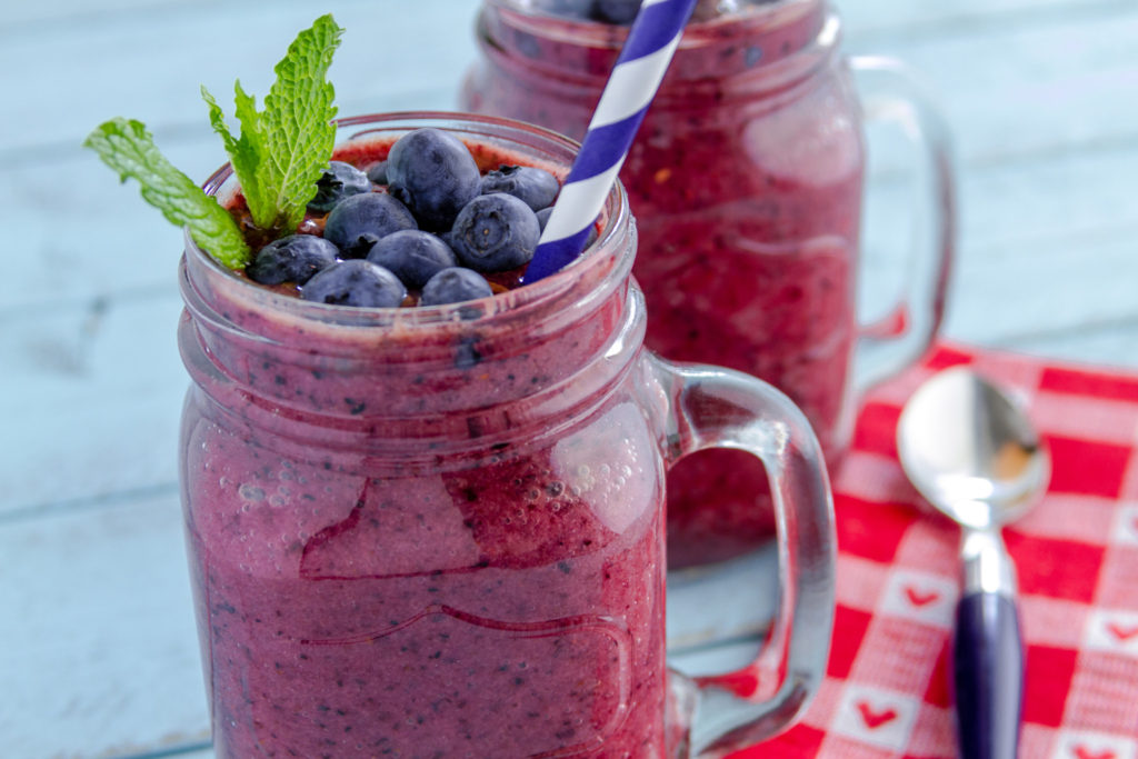 Close up of 2 Mason jars filled with blueberry and blackberry fresh fruit smoothie sitting on blue wood background with straws and heart napkin
