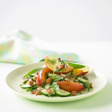 A fresh salad on a white plate features sliced peaches, cucumbers, mint leaves, and chopped almonds, garnished with a light dressing. A pastel-colored cloth with polka dots is in the blurred background.