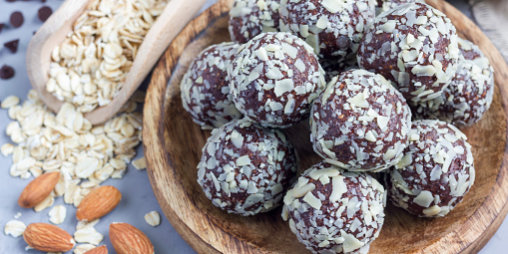 A wooden plate is filled with round chocolate energy bites, each coated with oats and a hint of pumpkin spice. Surrounding the plate are scattered almonds, chocolate chips, and a wooden scoop filled with rolled oats. The background is a light-colored surface.