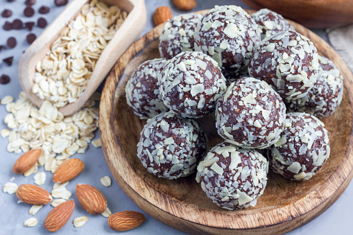 A wooden plate is filled with round chocolate energy bites, each coated with oats and a hint of pumpkin spice. Surrounding the plate are scattered almonds, chocolate chips, and a wooden scoop filled with rolled oats. The background is a light-colored surface.