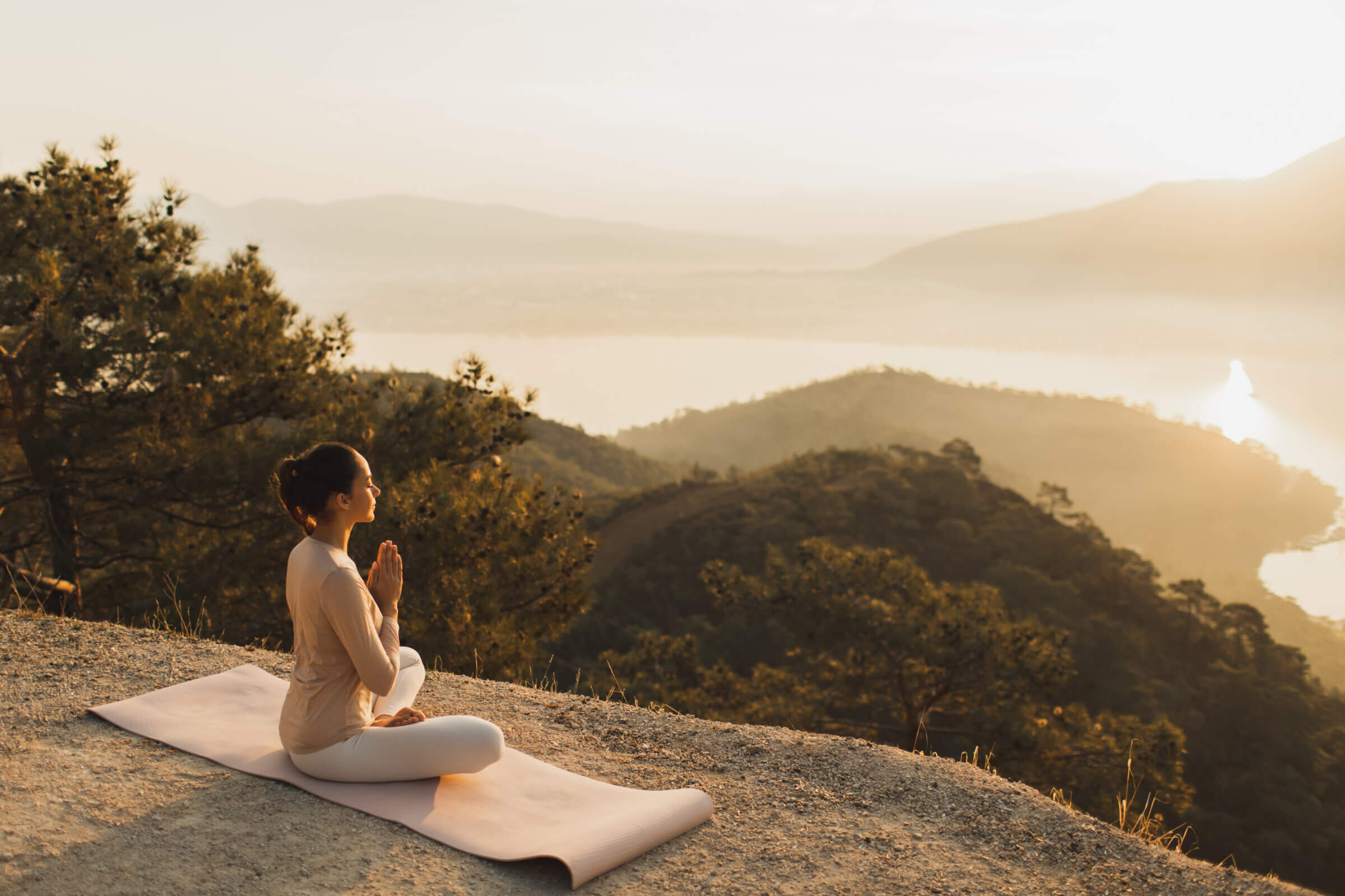 A person sits in a meditative pose on a mat atop a hill at sunset, overlooking a forested valley and distant lake.