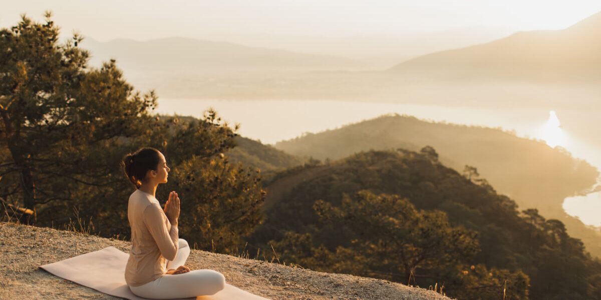 A person sits in a meditative pose on a mat atop a hill at sunset, overlooking a forested valley and distant lake.