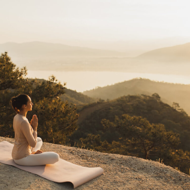 A person sits in a meditative pose on a mat atop a hill at sunset, overlooking a forested valley and distant lake.
