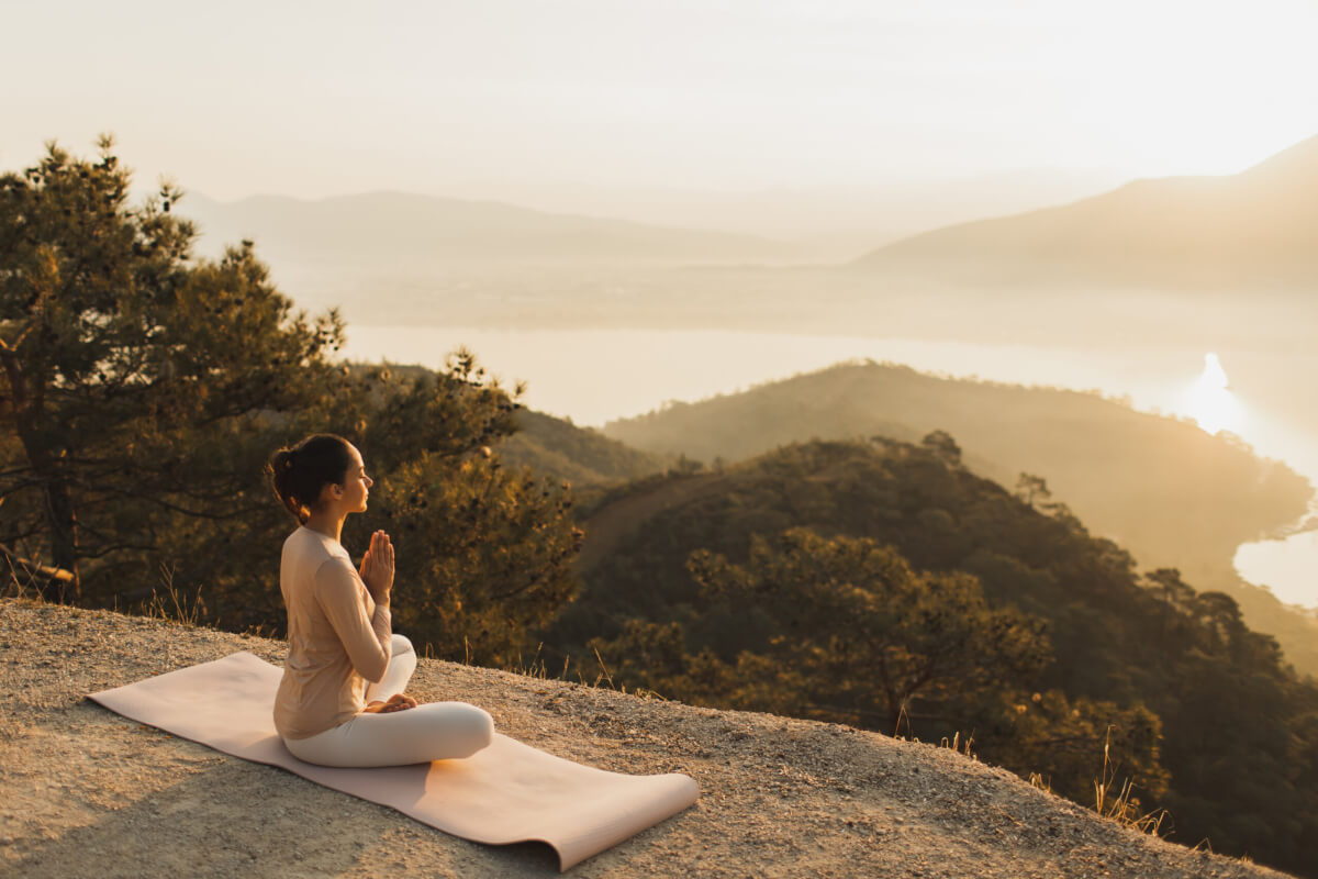 A person sits in a meditative pose on a mat atop a hill at sunset, overlooking a forested valley and distant lake.