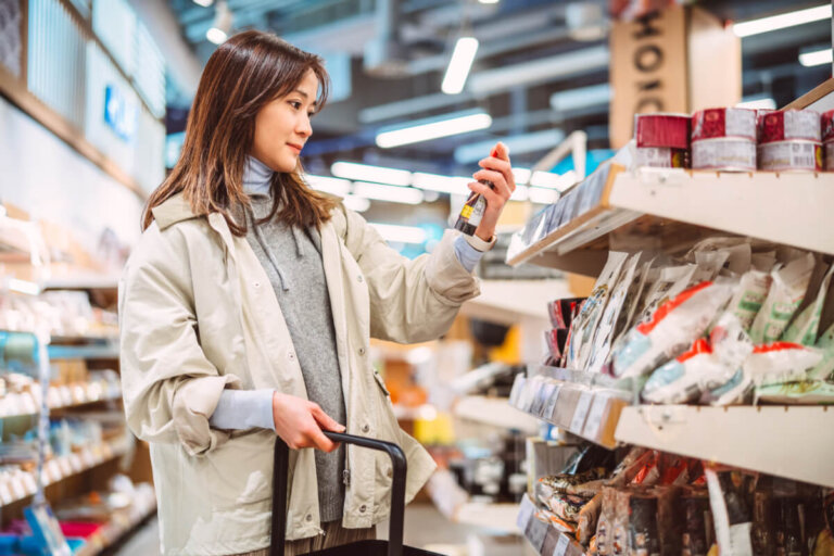 A woman in a light jacket stands in a supermarket aisle, holding a bottle and looking at it beside her shopping cart.