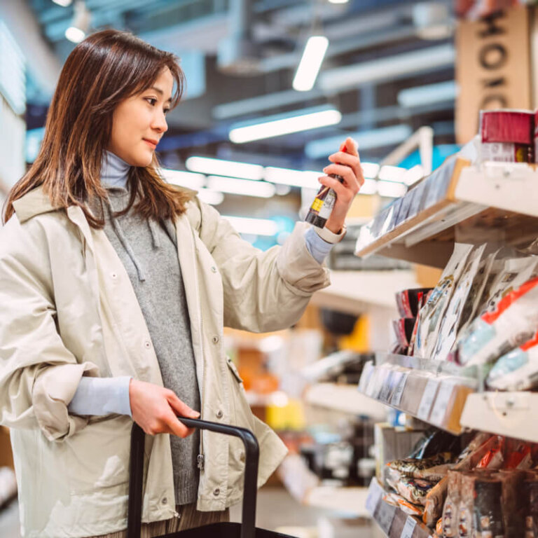 A woman in a light jacket stands in a supermarket aisle, holding a bottle and looking at it beside her shopping cart.