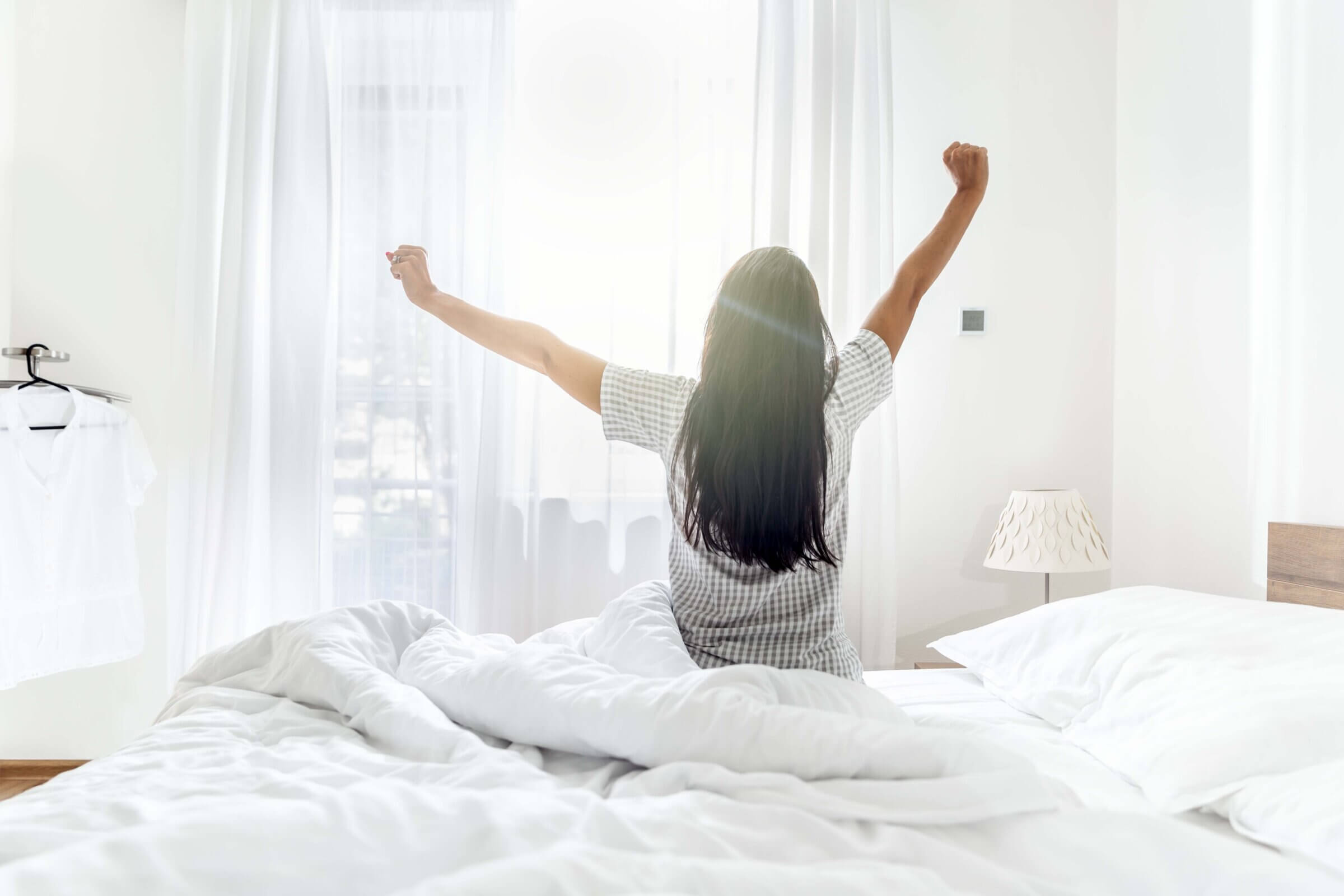Person sitting up in a white bed with arms raised, facing a sunlit window in a bright bedroom.