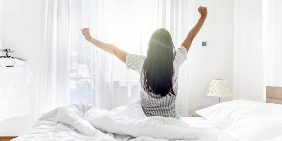 Person sitting up in a white bed with arms raised, facing a sunlit window in a bright bedroom.