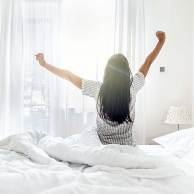 Person sitting up in a white bed with arms raised, facing a sunlit window in a bright bedroom.