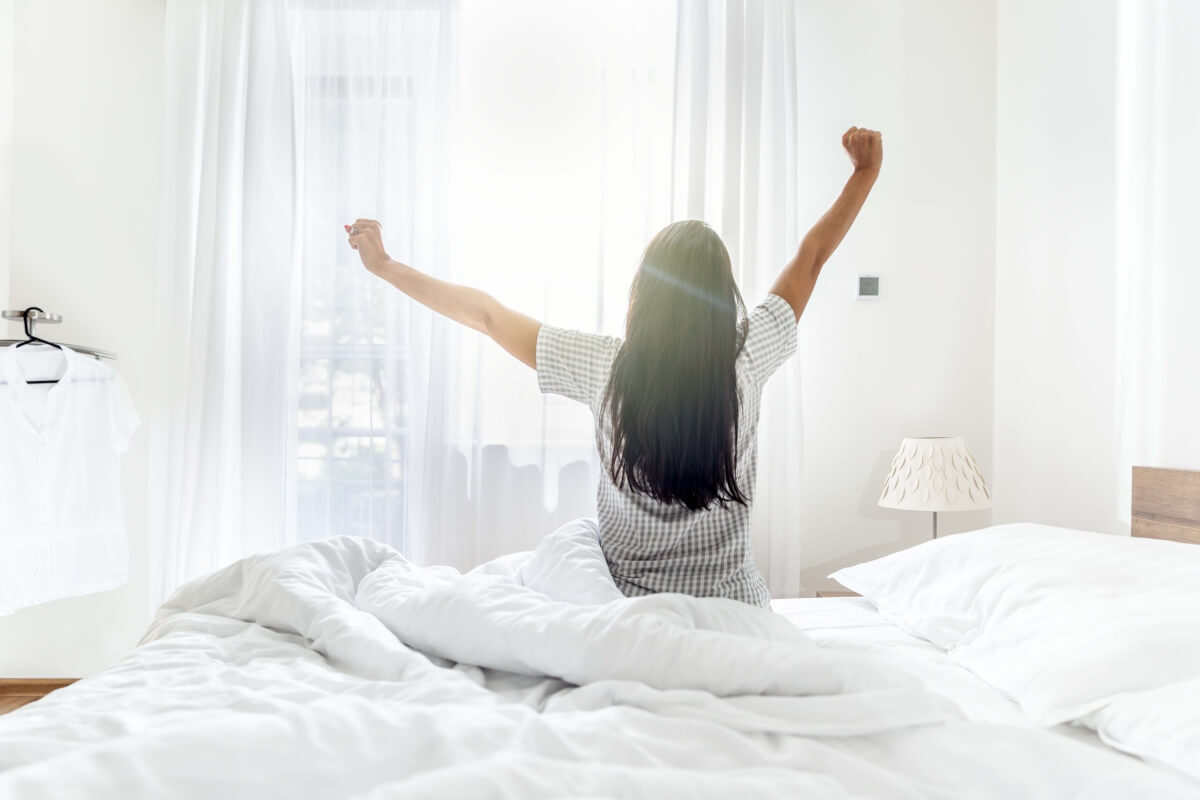 Person sitting up in a white bed with arms raised, facing a sunlit window in a bright bedroom.