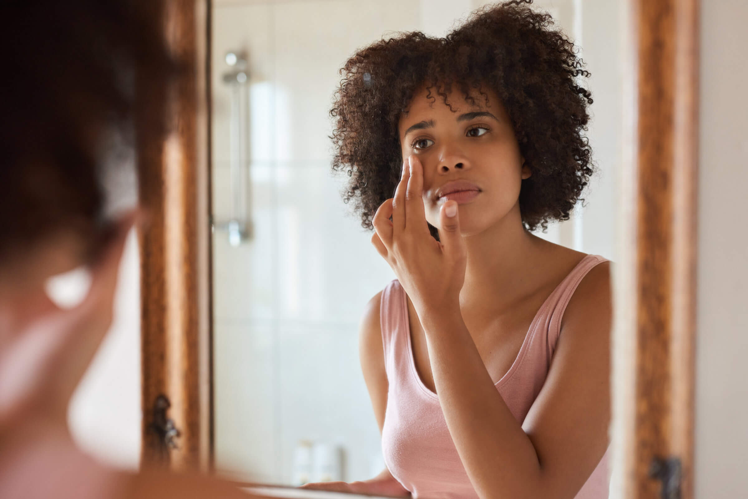 Woman with curly hair applying makeup in front of a bathroom mirror.
