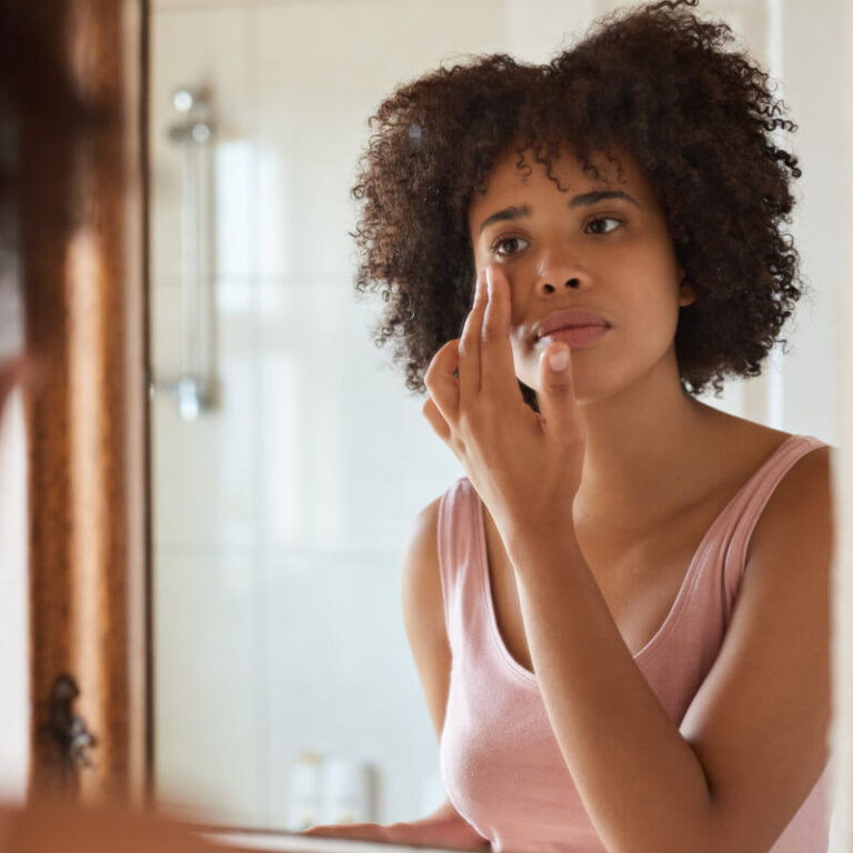 Woman with curly hair applying makeup in front of a bathroom mirror.