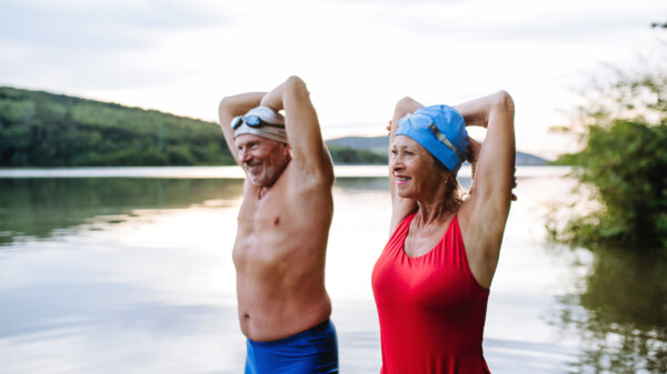 Smiling older couple standing in a lake with their arms raised behind their heads, wearing swim caps and swimsuits.