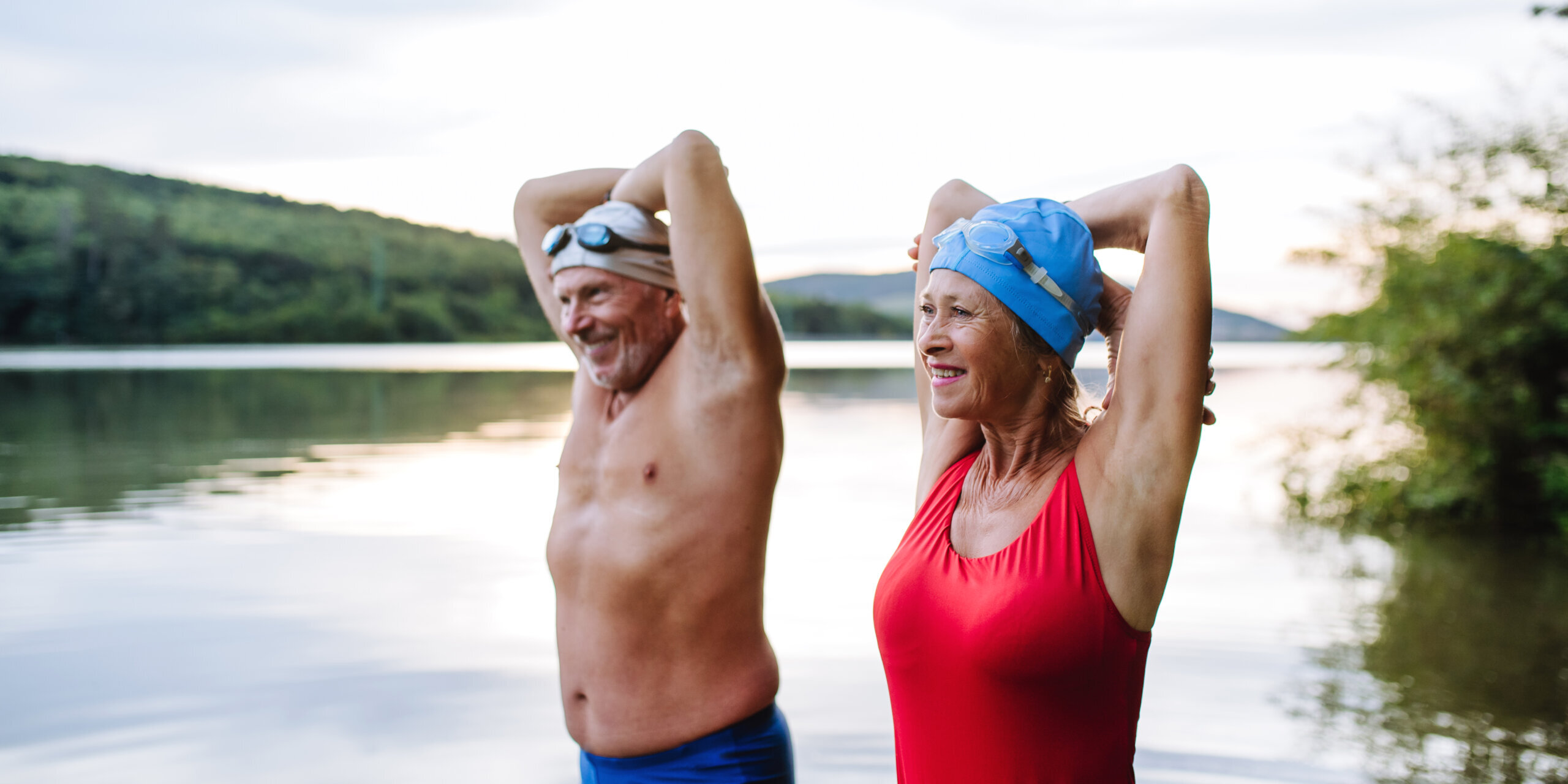 Senior couple has a shared hobby, swimming in lake during cold autumn morning. Stretching before going into water. Smiling older couple standing in a lake with their arms raised behind their heads, wearing swim caps and swimsuits.
