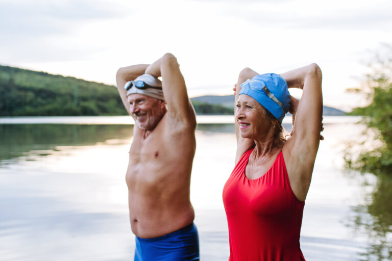Smiling older couple standing in a lake with their arms raised behind their heads, wearing swim caps and swimsuits.