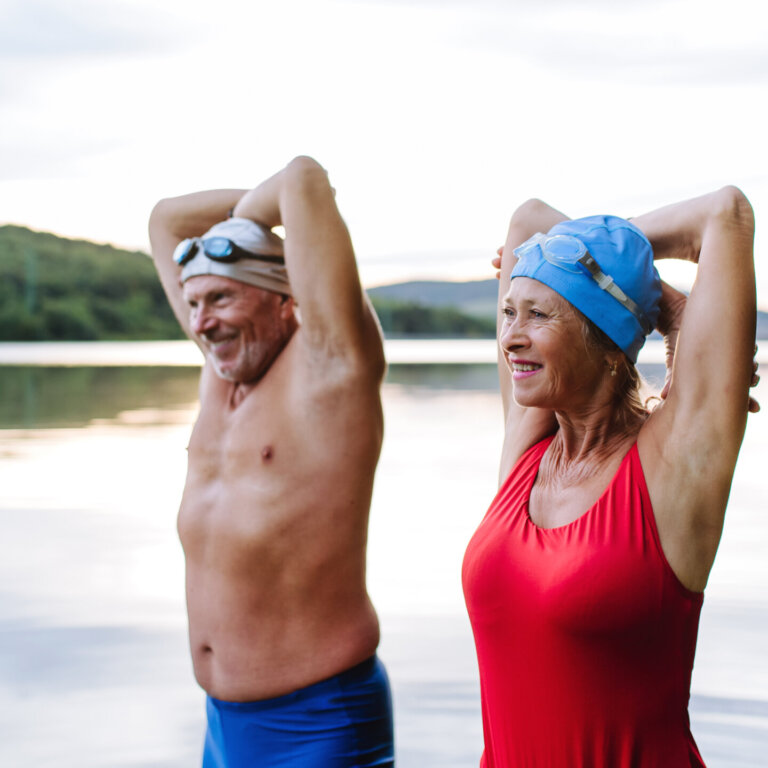Smiling older couple standing in a lake with their arms raised behind their heads, wearing swim caps and swimsuits.