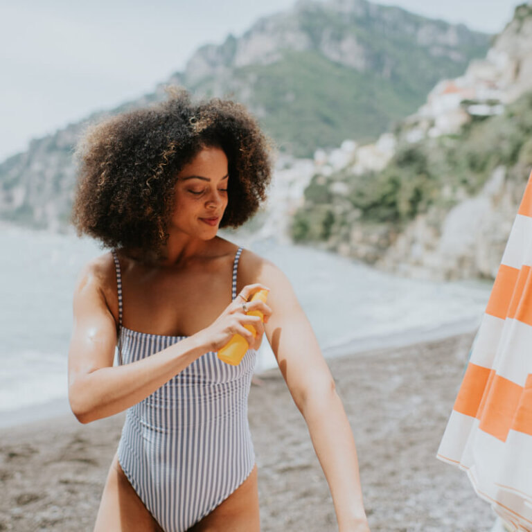 A woman in a striped swimsuit applies sunscreen to her arm on a beach beside an orange-and-white striped umbrella.