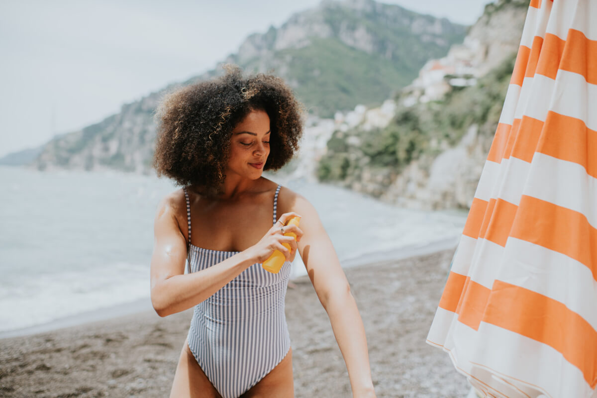 A woman in a striped swimsuit applies sunscreen to her arm on a beach beside an orange-and-white striped umbrella.