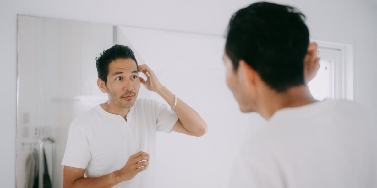 Man in a white shirt fixes his hair while looking at his reflection in a bathroom mirror.
