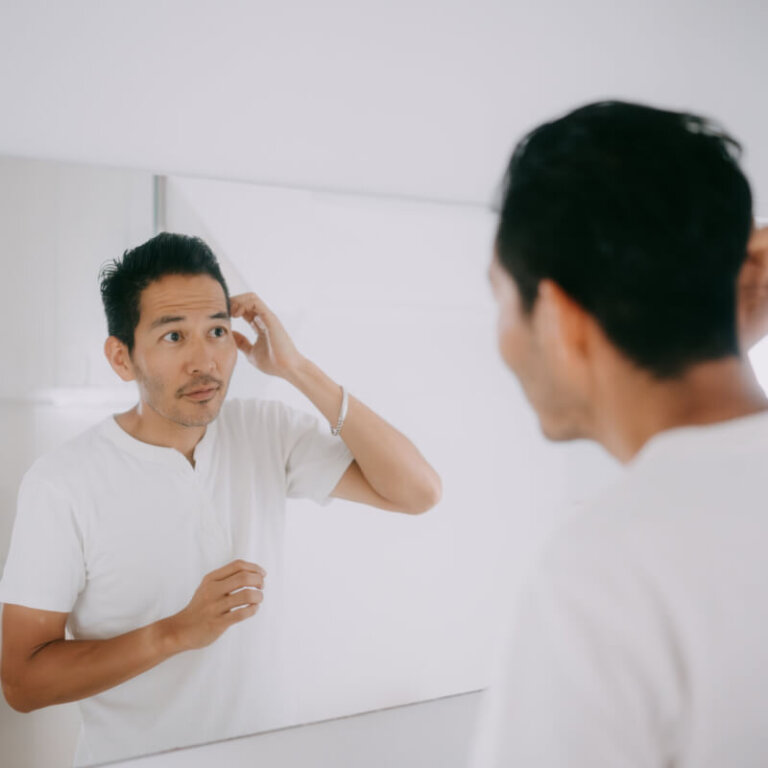Man in a white shirt fixes his hair while looking at his reflection in a bathroom mirror.