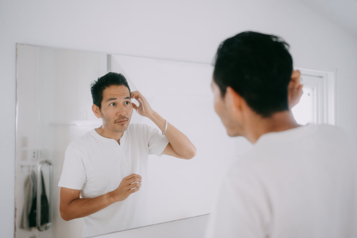 Man in a white shirt fixes his hair while looking at his reflection in a bathroom mirror.