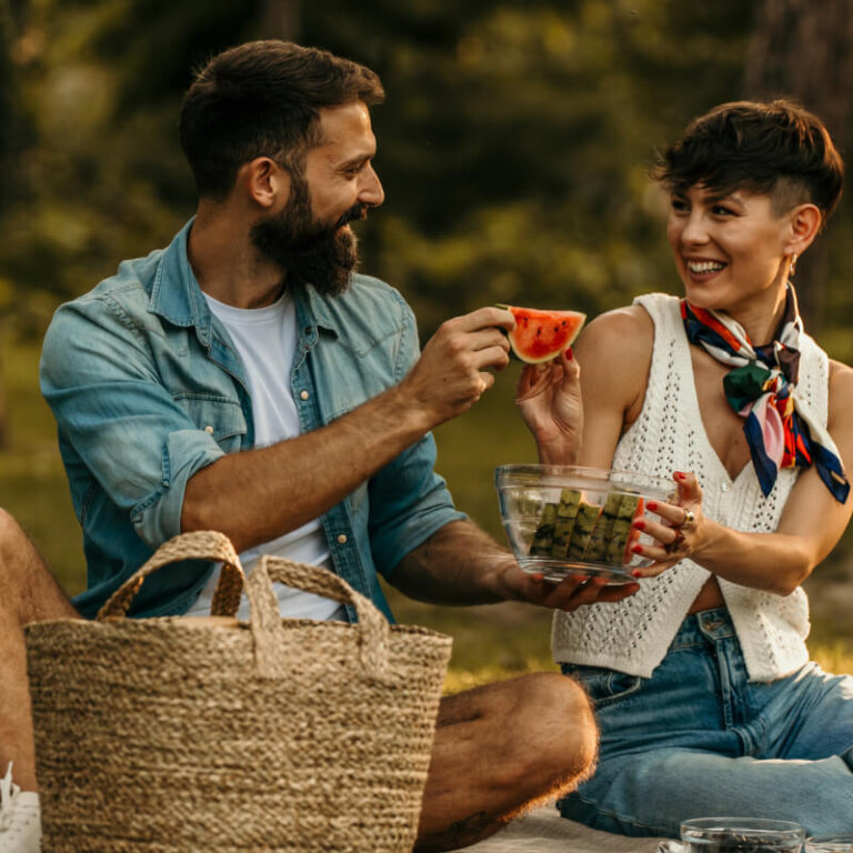 A man and a woman share a picnic on a park lawn, smiling as they pass a slice of watermelon.