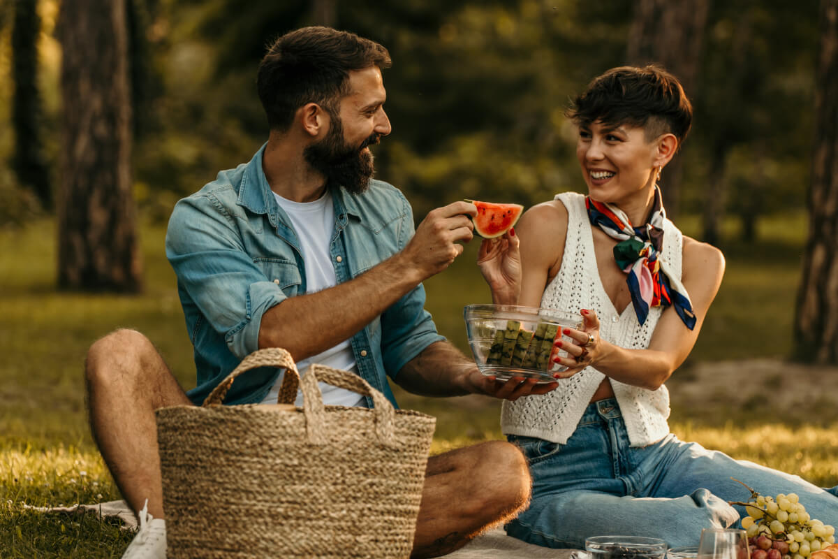 A man and a woman share a picnic on a park lawn, smiling as they pass a slice of watermelon.
