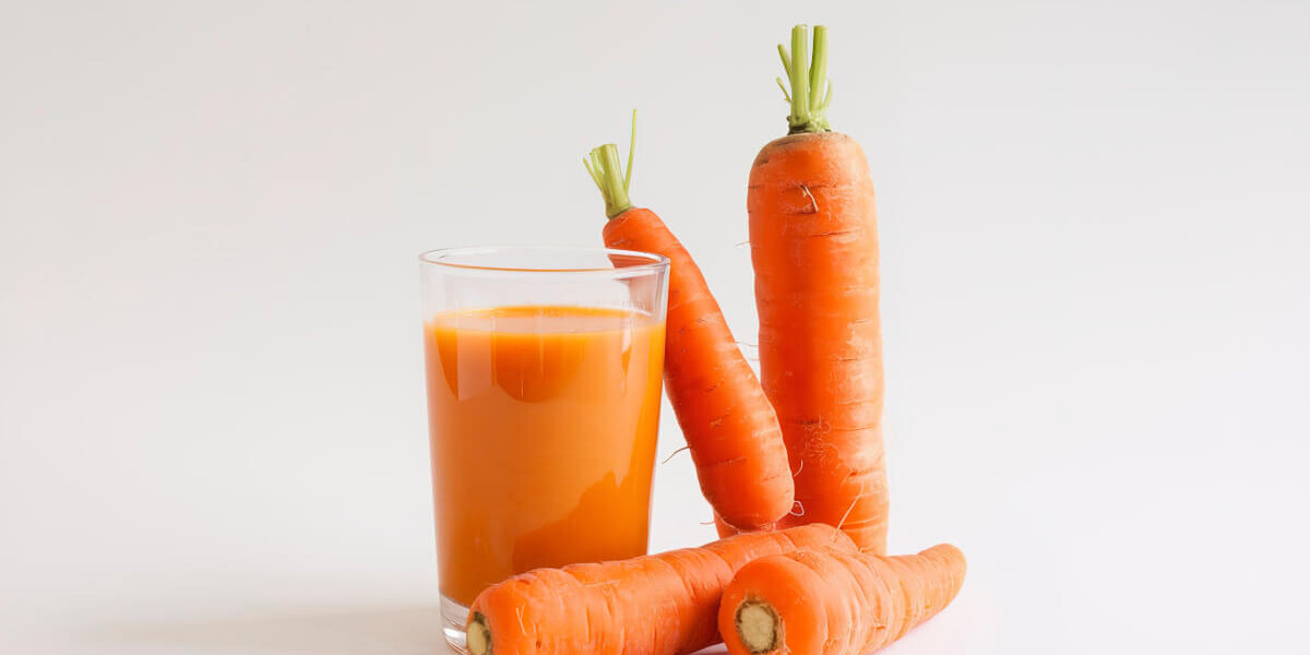 A glass of carrot juice sits beside several fresh carrots on a white background.