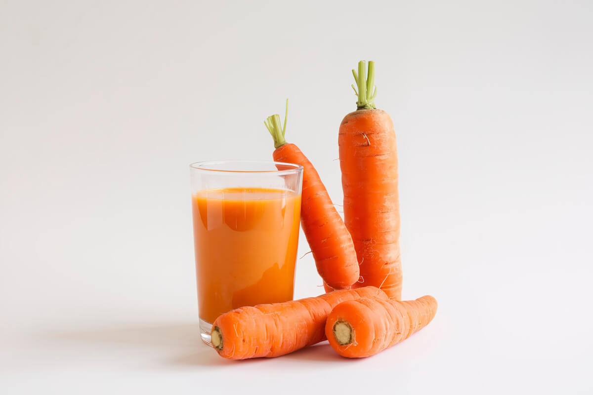 A glass of carrot juice sits beside several fresh carrots on a white background.