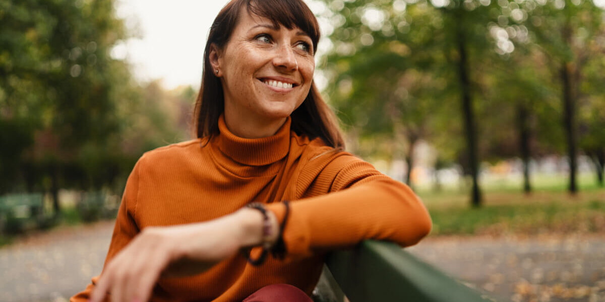 Smiling woman in an orange turtleneck leaning on a park bench with trees in the background.