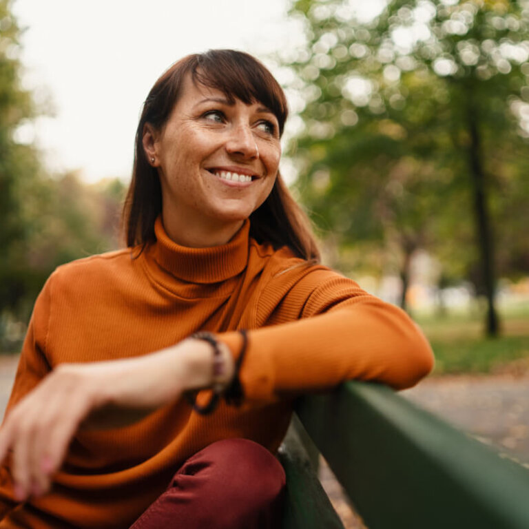 Smiling woman in an orange turtleneck leaning on a park bench with trees in the background.