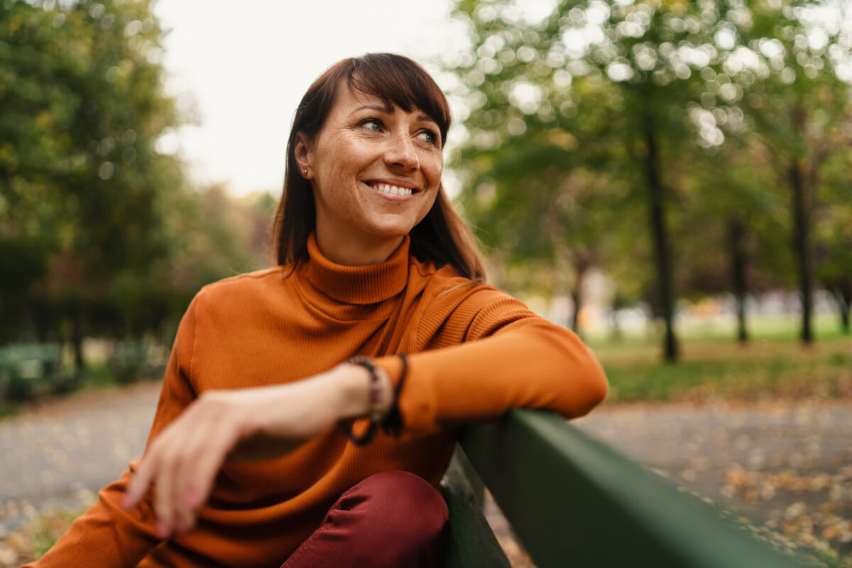 Smiling woman in an orange turtleneck leaning on a park bench with trees in the background.