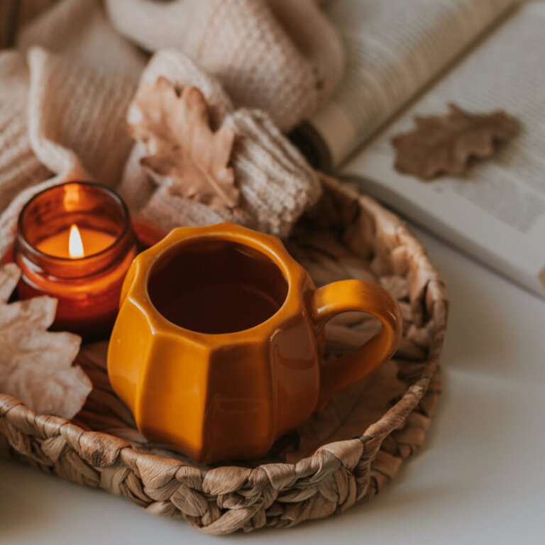 A cozy woven basket holds an orange ceramic mug, a small lit amber candle, soft knitted fabrics, and dried autumn leaves.