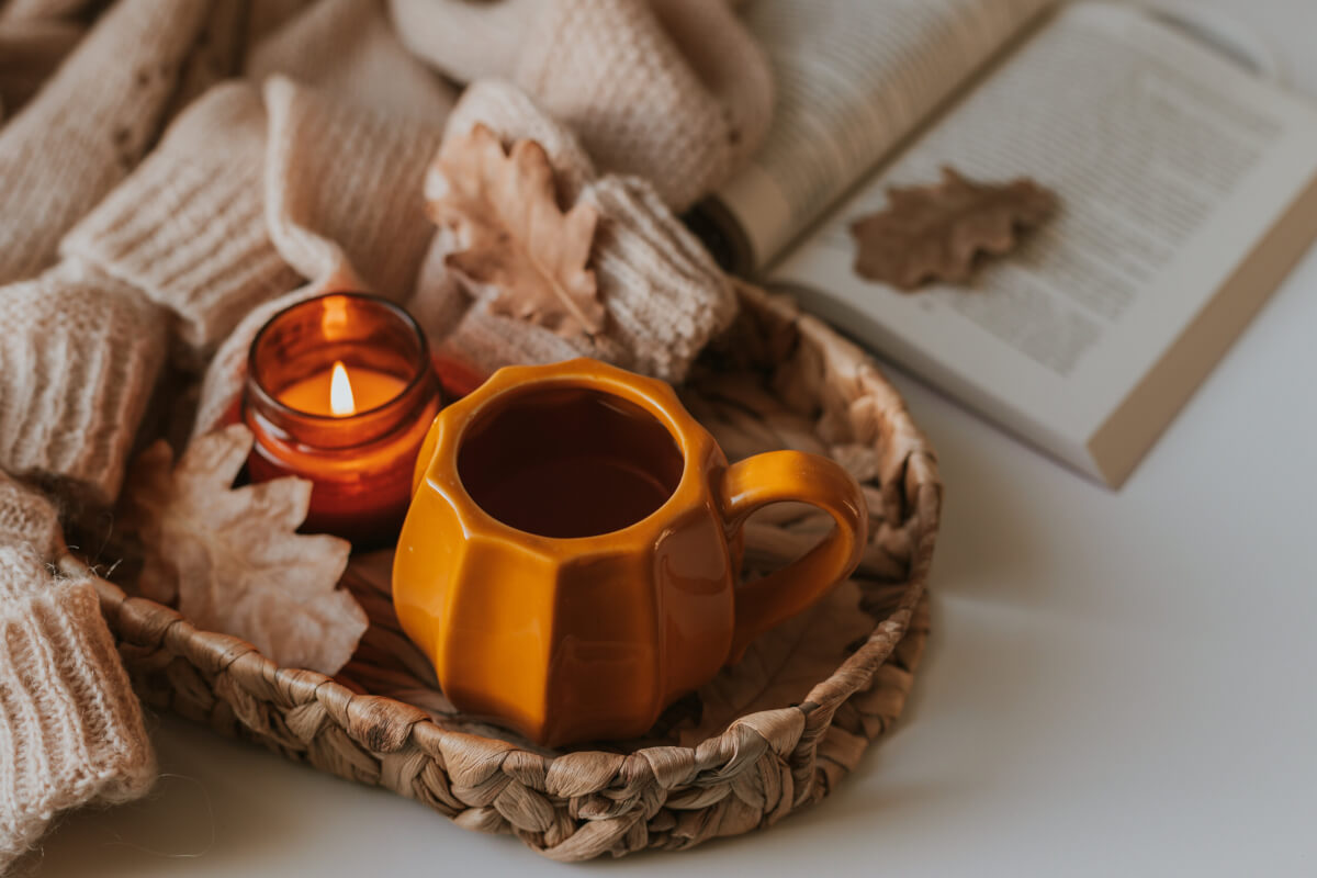 A cozy woven basket holds an orange ceramic mug, a small lit amber candle, soft knitted fabrics, and dried autumn leaves.