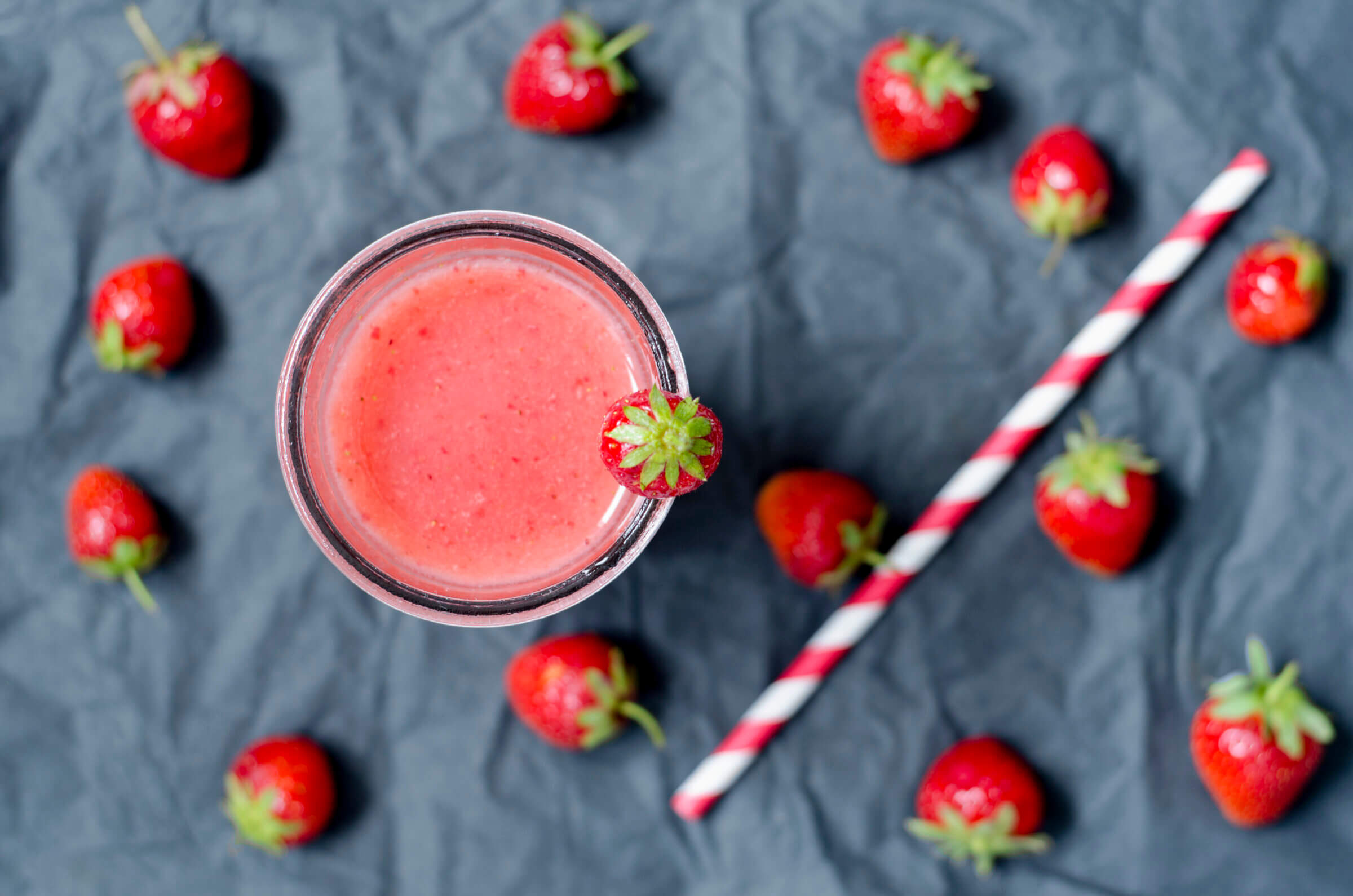 A glass of pink strawberry smoothie with a strawberry on the rim, surrounded by whole strawberries and a red-and-white striped straw on a dark surface.