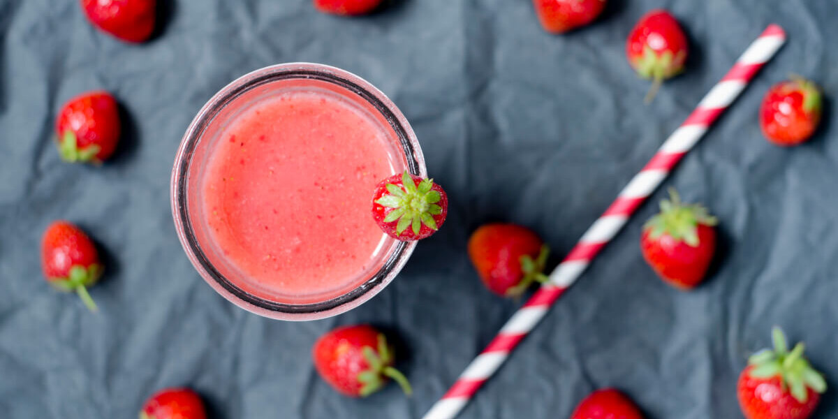A glass of pink strawberry smoothie with a strawberry on the rim, surrounded by whole strawberries and a red-and-white striped straw on a dark surface.