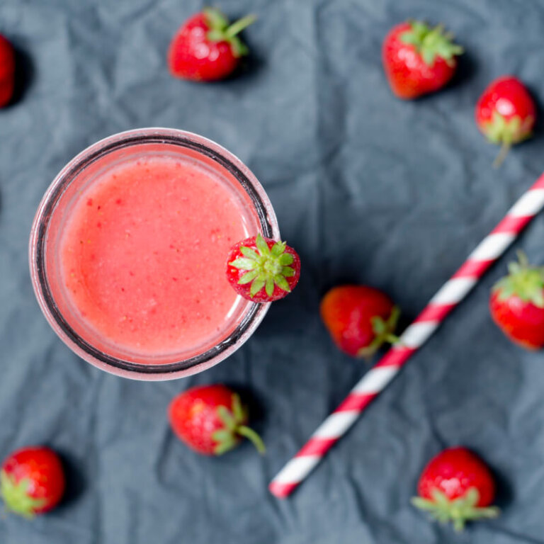 A glass of pink strawberry smoothie with a strawberry on the rim, surrounded by whole strawberries and a red-and-white striped straw on a dark surface.