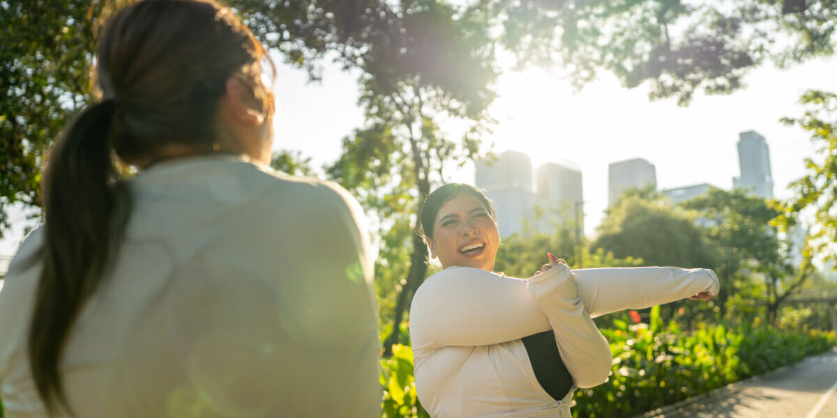 Two women in a sunlit park with trees and a city skyline in the background; one is smiling and stretching her arms while the other has her back to the camera.