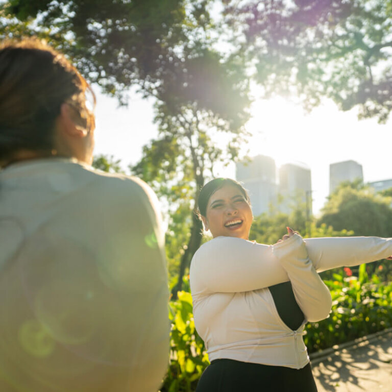 Two women in a sunlit park with trees and a city skyline in the background; one is smiling and stretching her arms while the other has her back to the camera.
