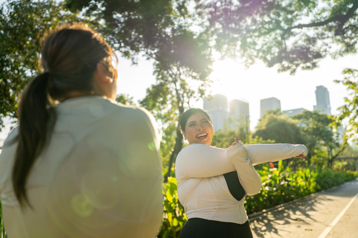 Two women in a sunlit park with trees and a city skyline in the background; one is smiling and stretching her arms while the other has her back to the camera.