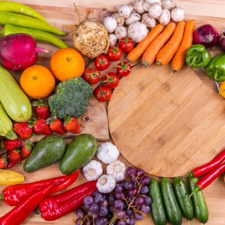 Colorful assortment of fresh fruits and vegetables arranged around a round wooden cutting board.