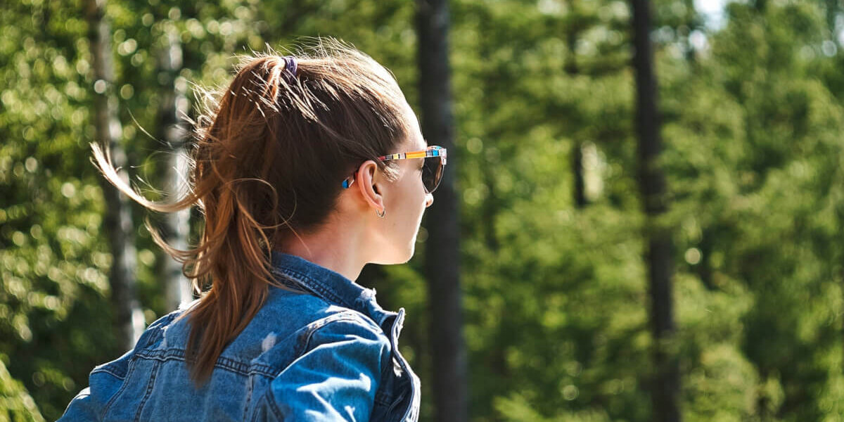 A person with a ponytail wearing a denim jacket and sunglasses stands in a sunlit forest.