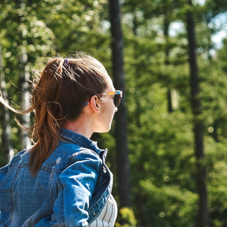 A person with a ponytail wearing a denim jacket and sunglasses stands in a sunlit forest.