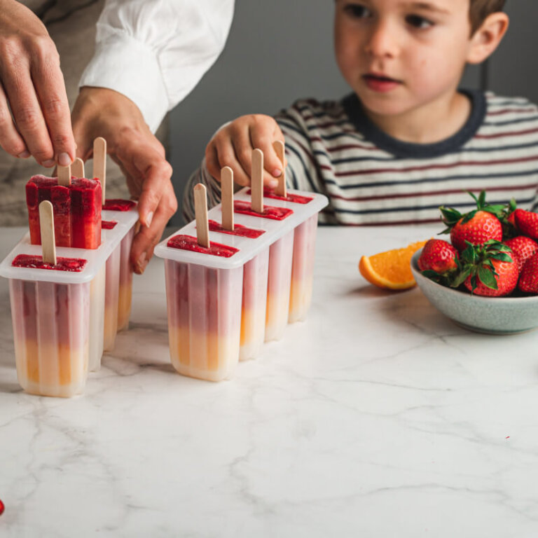 Child watches as an adult prepares layered fruit popsicles in rectangular molds on a marble countertop, with strawberries and oranges nearby.