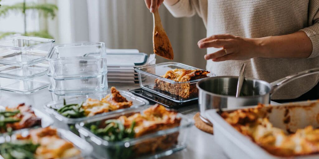 Person preparing and portioning meals into glass containers on a kitchen counter. The image shows a variety of dishes, including lasagna and green beans, being carefully arranged in meal prep containers, highlighting the organized and healthy approach to meal planning.
