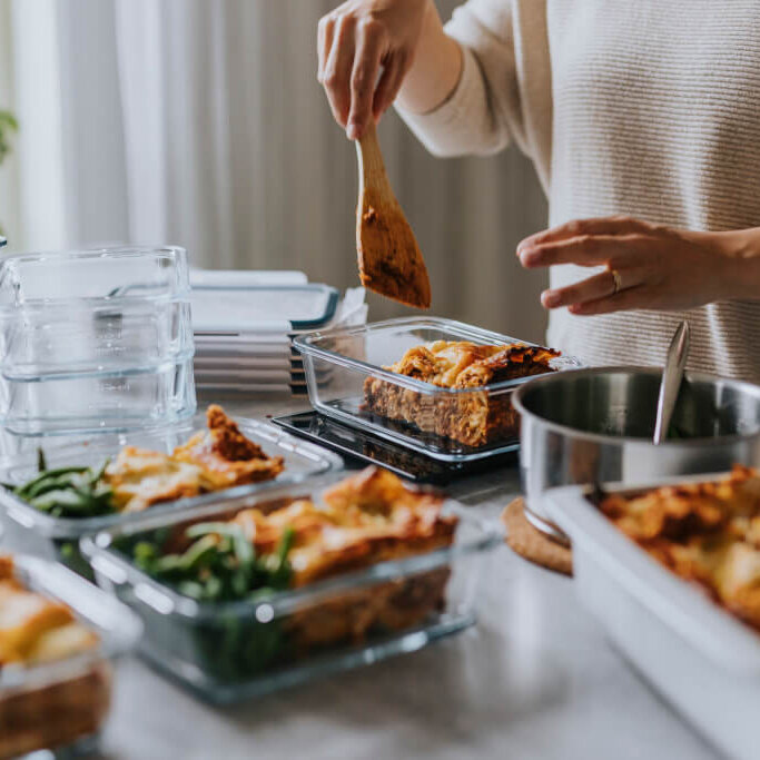 Person preparing and portioning meals into glass containers on a kitchen counter. The image shows a variety of dishes, including lasagna and green beans, being carefully arranged in meal prep containers, highlighting the organized and healthy approach to meal planning.