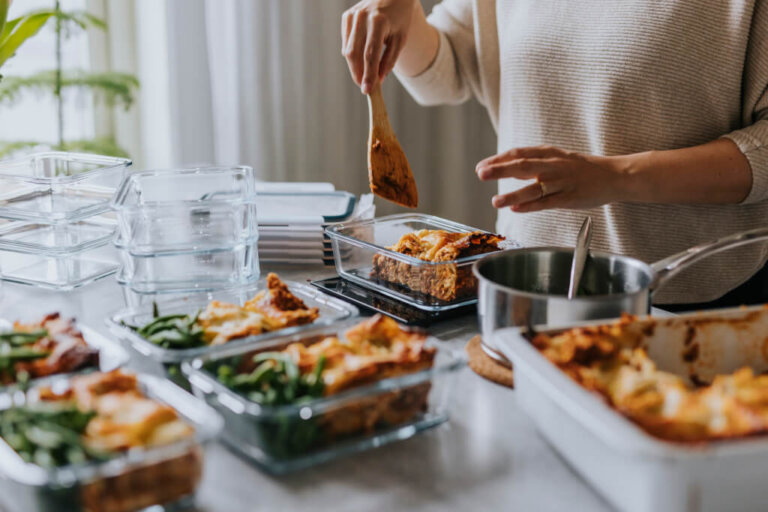 Person preparing and portioning meals into glass containers on a kitchen counter. The image shows a variety of dishes, including lasagna and green beans, being carefully arranged in meal prep containers, highlighting the organized and healthy approach to meal planning.