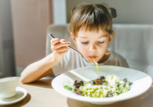 Young child happily eating a bowl of pesto pasta, with green sauce visible on the noodles, illustrating a creative way to incorporate more vegetables into meals. 