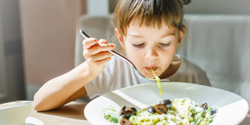 Young child happily eating a bowl of pesto pasta, with green sauce visible on the noodles, illustrating a creative way to incorporate more vegetables into meals. "Young child happily eating a bowl of pesto pasta, with green sauce visible on the noodles, illustrating a creative way to incorporate more vegetables into meals."
