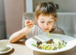 Young child happily eating a bowl of pesto pasta, with green sauce visible on the noodles, illustrating a creative way to incorporate more vegetables into meals. "Young child happily eating a bowl of pesto pasta, with green sauce visible on the noodles, illustrating a creative way to incorporate more vegetables into meals."